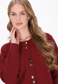 Cardigan tricotat roșu cu un finisaj texturat, având un guler rotund și cinci butoane de culoare aurie pe față.