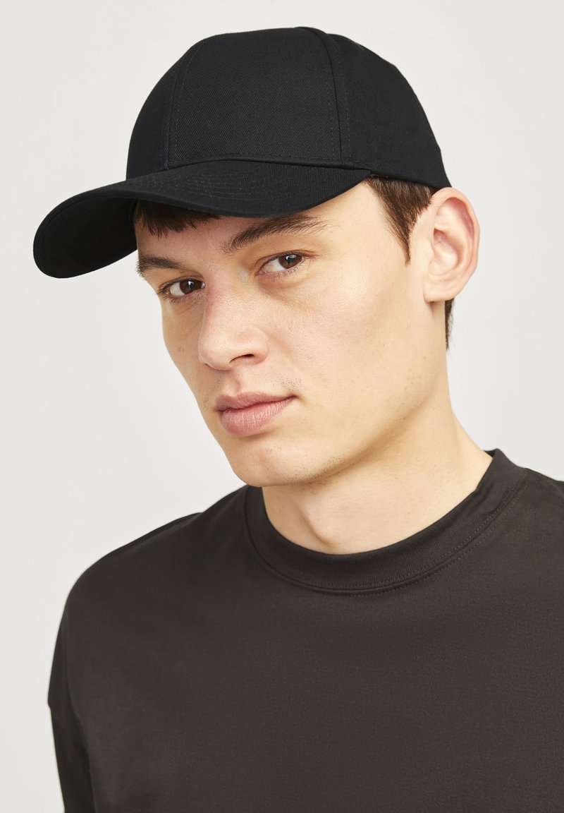 Căciulă de baseball neagră din material textil, cu un bor mai curbat, design cu șase panouri și cusături întărite. Purtați de un model cu o expresie neutră.