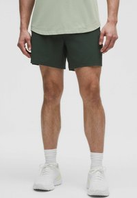 Dunkelgrüne Sportshorts mit elastischem Bund, kombiniert mit einem hellgrünen Shirt und weißen Sneakers. Der Stoff wirkt leicht und glatt.