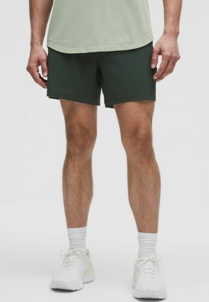 Mörkgröna sportshorts med en platt midja, tillsammans med en ljusgrön skjorta och vita sneakers. Tyget verkar lätt och mjukt.