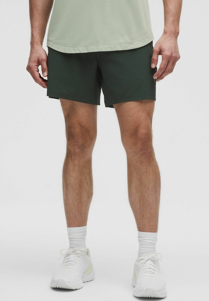 Dunkelgrüne Sportshorts mit elastischem Bund, kombiniert mit einem hellgrünen Shirt und weißen Sneakers. Der Stoff wirkt leicht und glatt.