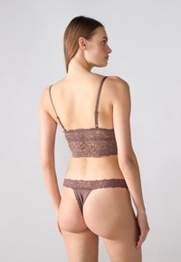 Smeđi čipkasti bralette s podesivim naramenicama u paru s odgovarajućim tangama. Čipka ima cvjetni uzorak i mekan, glatki tekstur.