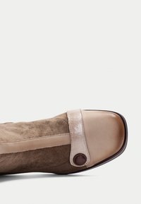 Zapato de ante marrón con una banda de piel beige en la parte delantera y un botón decorativo. Sección delantera suave y brillante con una forma redondeada.