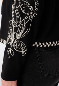 Zwarte stof met witte bloemenborduursels op de mouwen en zoom, gedragen met zwarte denim jeans met details van de achterzak.