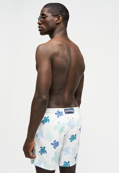 Άνδρας χωρίς πουκάμισο, φορώντας λευκά μαγιό με μοτίβα χελωνών σε μπλε και πράσινο, γυρισμένος ελαφρώς στη μία πλευρά, φορώντας γυαλιά ηλίου.