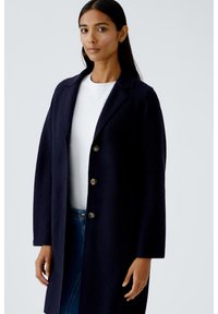 Marineblauer Wollmantel mit maßgeschneidertem Design, mit Reverskragen, langen Ärmeln und vier großen Knöpfen auf der Vorderseite. Glatte Textur und mittellanger Stil.