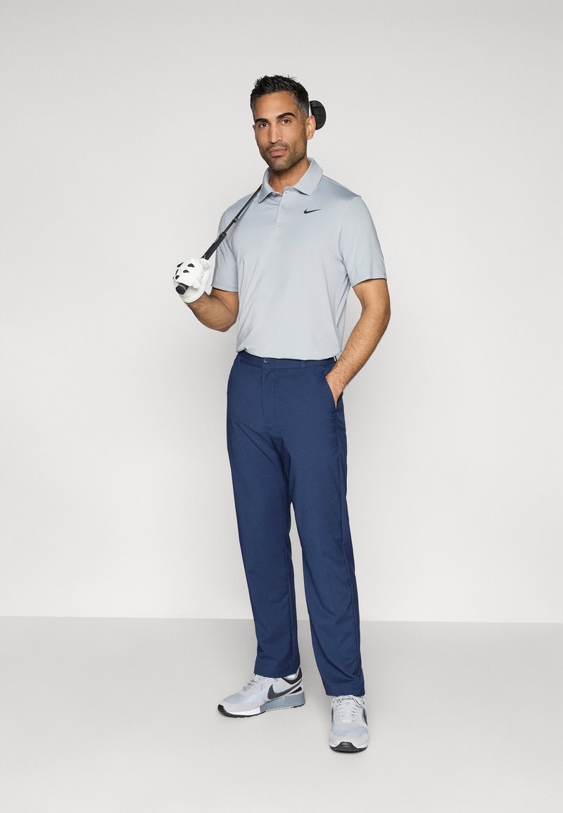 Graues Poloshirt, marineblaue Hose und graue Sportschuhe. Ein Golfschläger ruht auf der Schulter; der Golfer trägt einen weißen Handschuh an einer Hand.
