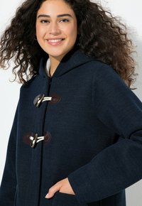 Tmavě modrý kapucí kabát s texturovaným povrchem, opatřený zapínáním na knoflíky ve tmavě hnědé a krémové barvě, bočními kapsami a volnějším střihem.