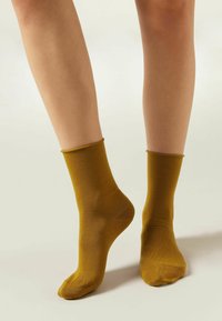 Calcetines de tobillo color mostaza, con textura suave, forma tubular, y con un puño acanalado. Se ajustan de cerca al tobillo sin adornos.