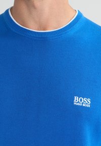 Niebieski sweter dzianinowy z okrągłym dekoltem, z białym prążkowanym wykończeniem i małym białym logo na lewej piersi. Gładka faktura.
