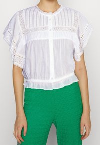 Blusa bianca leggera con maniche corte a volant, dettagli in pizzo elaborati, chiusura con bottoni e orlo arricciato, abbinata a pantaloni verdi texturizzati.