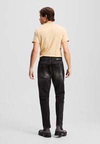 Jeans de mezclilla negra con un acabado desgastado, diseño de cinco bolsillos y una etiqueta en la cinturilla. Combinados con una camiseta amarilla clara.