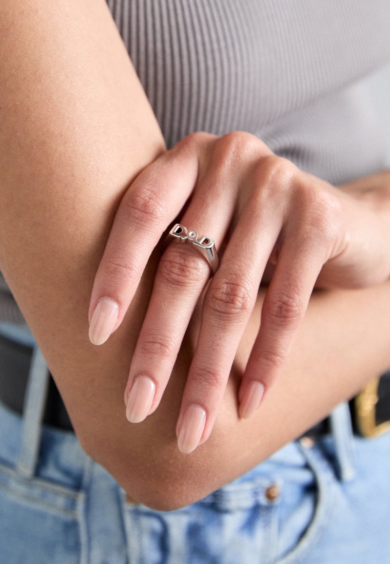 Anello d'argento con un design moderno e dettagli a forma di "D", indossato su una mano con unghie curate e smaltate di rosa chiaro, su uno sfondo neutro.