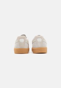 Ett par beiga mockasneakers med gummisulor som visas från baksidan mot en vit bakgrund.