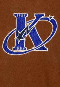 Tessuto marrone con un logo ricamato blu che presenta una "K" stilizzata e una stella, accompagnato da "1987" e "IKKS" in bianco.