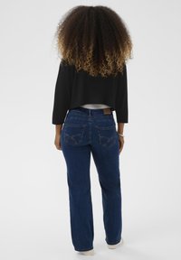 Frau mit lockigem Ombre-Haar, die von hinten steht, trägt dunkle Jeans, ein schwarzes Crop-Top und weiße Schuhe vor einem einfarbigen Hintergrund.