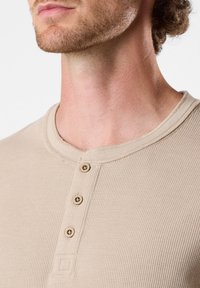 Beige geripptes Henley-Shirt mit rundem Ausschnitt, drei Holzknöpfen und dezenten Texturdetails auf dem Stoff.