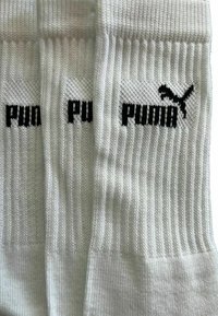 Drei weiße gerippte Socken flach ausgelegt, jede zeigt teilweise bis vollständig das schwarze Puma-Logo mit dem springenden Puma an der oberen Vorderseite.