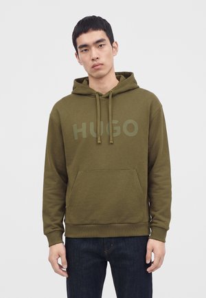 Νέος άνδρας που φοράει ελιάς πράσινο hoodie με μπροστινή τσέπη και κείμενο "HUGO", συνδυασμένο με σκούρο μπλε τζιν, στέκεται μπροστά σε απλό φόντο.
