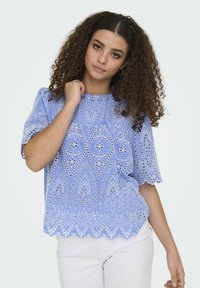 Blusa ricamata azzurro chiaro con scollatura rotonda, maniche puff corte e orlo a scallop. Presenta motivi floreali e paisley.