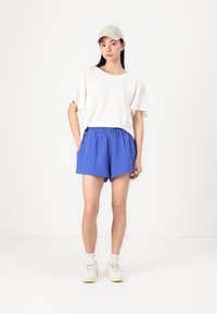 Lys beige oversized t-skjorte og blå linne shorts med snøring, kombinert med hvite sneakers og en brun caps, stående foran en nøytral bakgrunn.