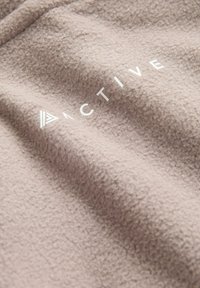 Fleece stof in gedempt beige met een zachte, gestructureerde oppervlakte. Bevat witte letters die "ACTIVE" spellen en een driehoekig accent.