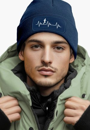 Navy knitted beanie with a textured pattern and a rectangular patch featuring a white heartbeat graphic. Worn with a green puffer jacket.