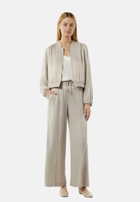 Beżowa satynowa kurtka bomberka z marszczonymi detalami, połączona z dopasowanymi spodniami o szerokich nogawkach. Stylizacja ma luźny krój i elastyczny pasek w talii.