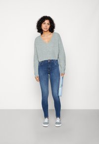 Suéter de punto gris claro con cuello en V, combinado con jeans ajustados azul oscuro. Sosteniendo un pequeño bolso cruzado azul claro, lleva zapatillas grises.