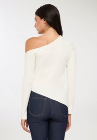 Kobieta z długimi ciemnymi włosami, ubrana w białą asymetryczną bluzkę bez ramion i ciemnoniebieskie dopasowane dżinsy, stoi tyłem na jednolitym tle.