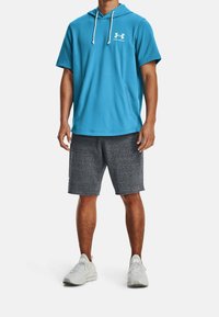 Tyrkysová krátkosrstá košile s kapucí má přední kapsu; je kombinována se šedými texturovanými šortkami. Bílé tenisky dotvářejí sportovní vzhled.