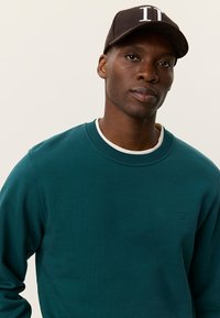 Τιρκουάζ φούτερ με στρογγυλή λαιμόκοψη και υφασμάτινη υφή, με διακριτικό λογότυπο στο στήθος. Μαύρο καπέλο με λευκά γράμματα στο μπροστινό μέρος.