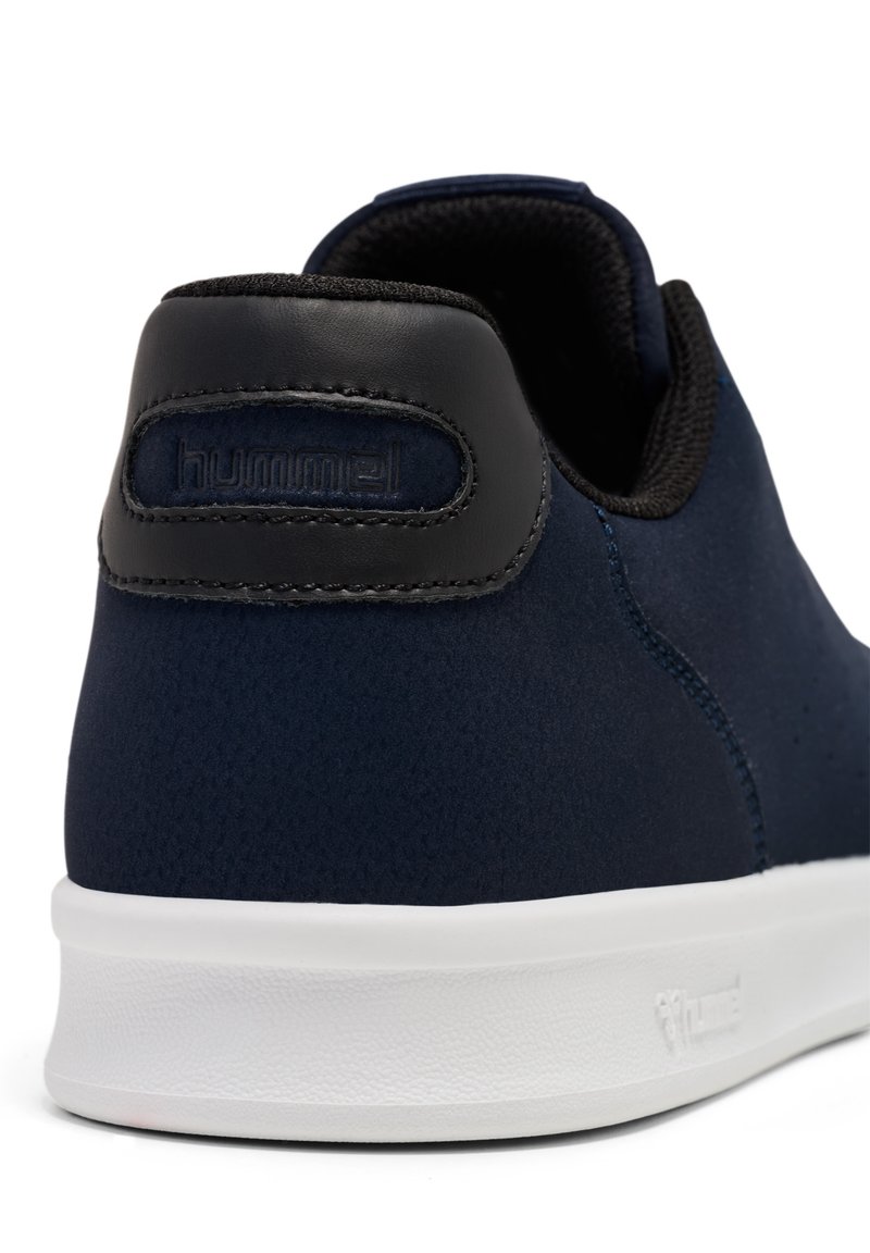 Zapatilla oscura azul con parte superior de tejido texturizado y acentos de cuero negro. Presenta una suela de goma blanca y una pestaña trasera con la marca.