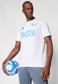 Hvid t-shirt med blå "Messi" tekst, parret med grå sportbukser med orange detaljer. En multicolor fodbold i hånden.