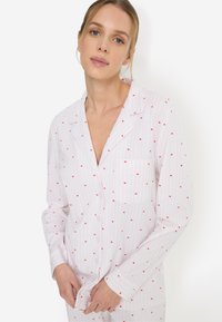 Σετ πυτζάμες με μακριά μανίκια από απαλό βαμβάκι, με ροζ και λευκές ρίγες και μικρές κόκκινες καρδιές. Περιλαμβάνει εμπρόσθιο κούμπωμα με κουμπιά και τσέπη στο στήθος.