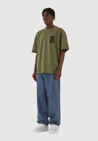 Oliven grøn oversized T-shirt med en sort grafik på brystet, parret med lysblå baggy jeans og hvide sneakers.