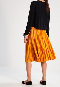 Blusa nera a maniche lunghe con aperture sulle spalle abbinata a una gonna midi plissettata arancione; il modello indossa ballerine nere.