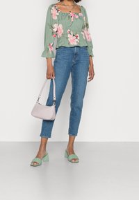 Donna che indossa una blusa floreale verde, jeans blu, sandali con tacco intrecciati verdi e tiene una borsa rosa chiaro su uno sfondo chiaro.