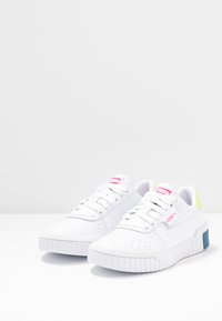 White sneakers with a textured leather upper, featuring perforations, pink and yellow accents, a thick sole, and a distinctive brand logo.
