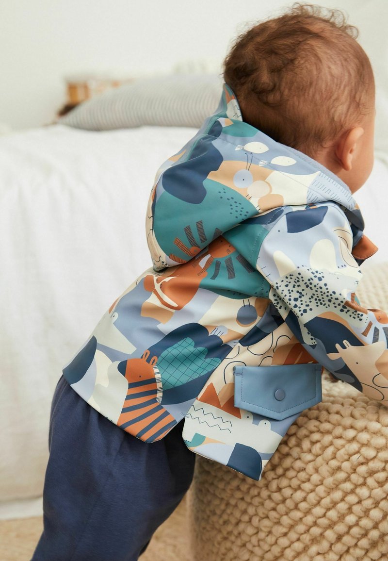 Giacca impermeabile colorata e fantasiosa con animali e forme, dotata di un cappuccio blu e una tasca frontale. Indossata sopra pantaloni scuri.