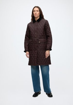 Femme debout portant un long manteau matelassé marron foncé avec col noir, jean bleu et chaussures noires sur un fond clair uni.