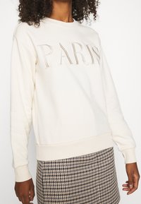 Felpa crema con "PARIGI" ricamato in beige chiaro. Presenta colletto e polsini a costine. Abbinata a una gonna a motivo pied de poule.