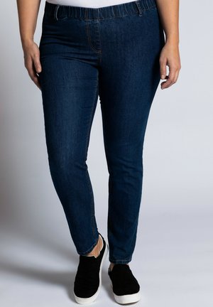 Donkerblauwe spijkerbroek met een elastische tailleband, slim fit en subtiele stikdetails, gecombineerd met zwarte instappers.