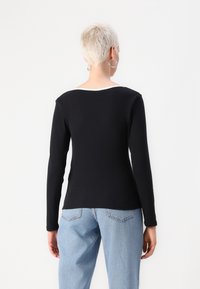 Svart langermet topp med hvit halslinning, tettsittende design, ribbet tekstur. Modellen har på seg lyseblå baggy jeans, sett fra baksiden.