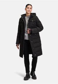Sort polstret lang frakke med hætte, lynlås lukning, forlommer og tommelhulsmuffer, båret med en grå sweater og sorte bukser.