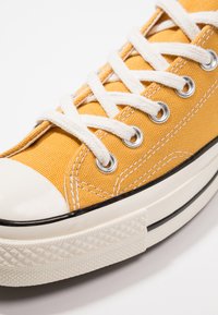 Gul canvas sneakers med hvite lisser, gummitåhette og kontrastfarge svart stripe langs sålen. Teksturet stoff og runde hull synlige.
