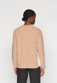 Beige teksturoitu huppari, jossa on raglan-hihat ja valkoiset sivupaneelit, esitelty takaa päin. Ominaisuuksiin kuuluu pyöreä pääntie ja pehmeä kangas.