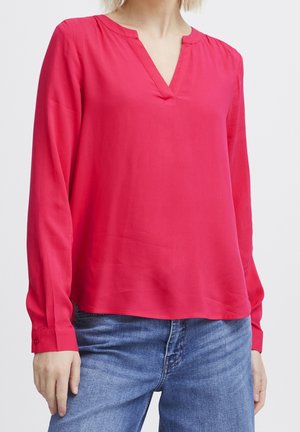 Kobieta w jasnoróżowej bluzce z długim rękawem i dekoltem w serek, w połączeniu z niebieskimi jeansami, stoi na jednolitym tle.