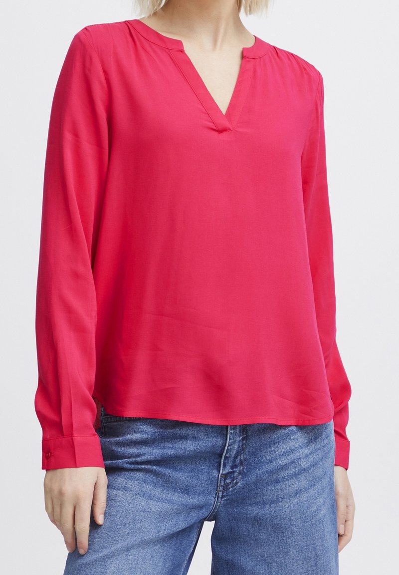 Femme portant un chemisier rose vif à manches longues avec un col en V, associé à un jean en denim bleu, debout devant un fond uni.
