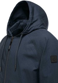 Navy hooded jacket with a lightweight, textured fabric. Features a zipper, adjustable drawstrings, and a small logo patch on the sleeve.